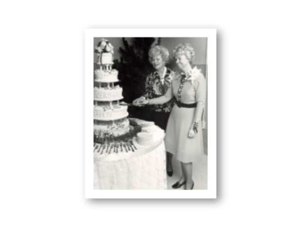 Two women standing next to a multi-tiered wedding cake on a white background