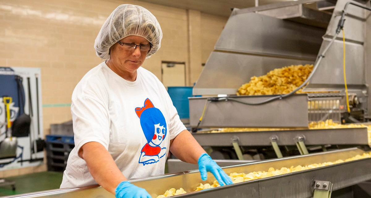 Person in a food processing facility wearing a hairnet and gloves, working with machinery.