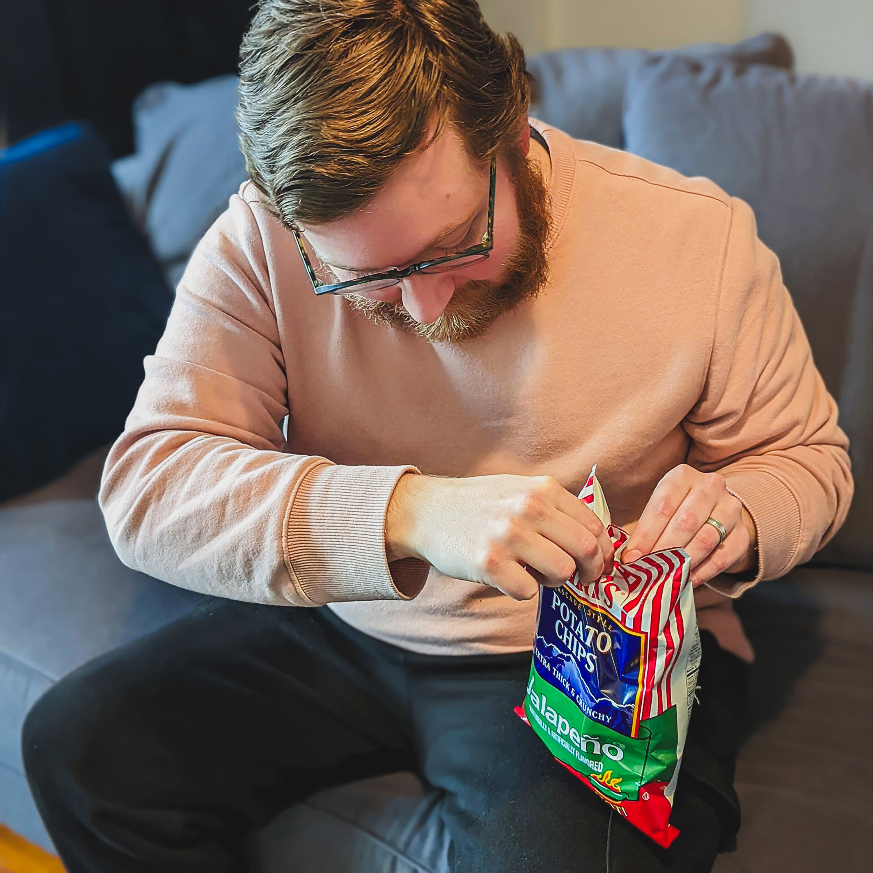 Man sitting on a couch opening a bag of potato chips.