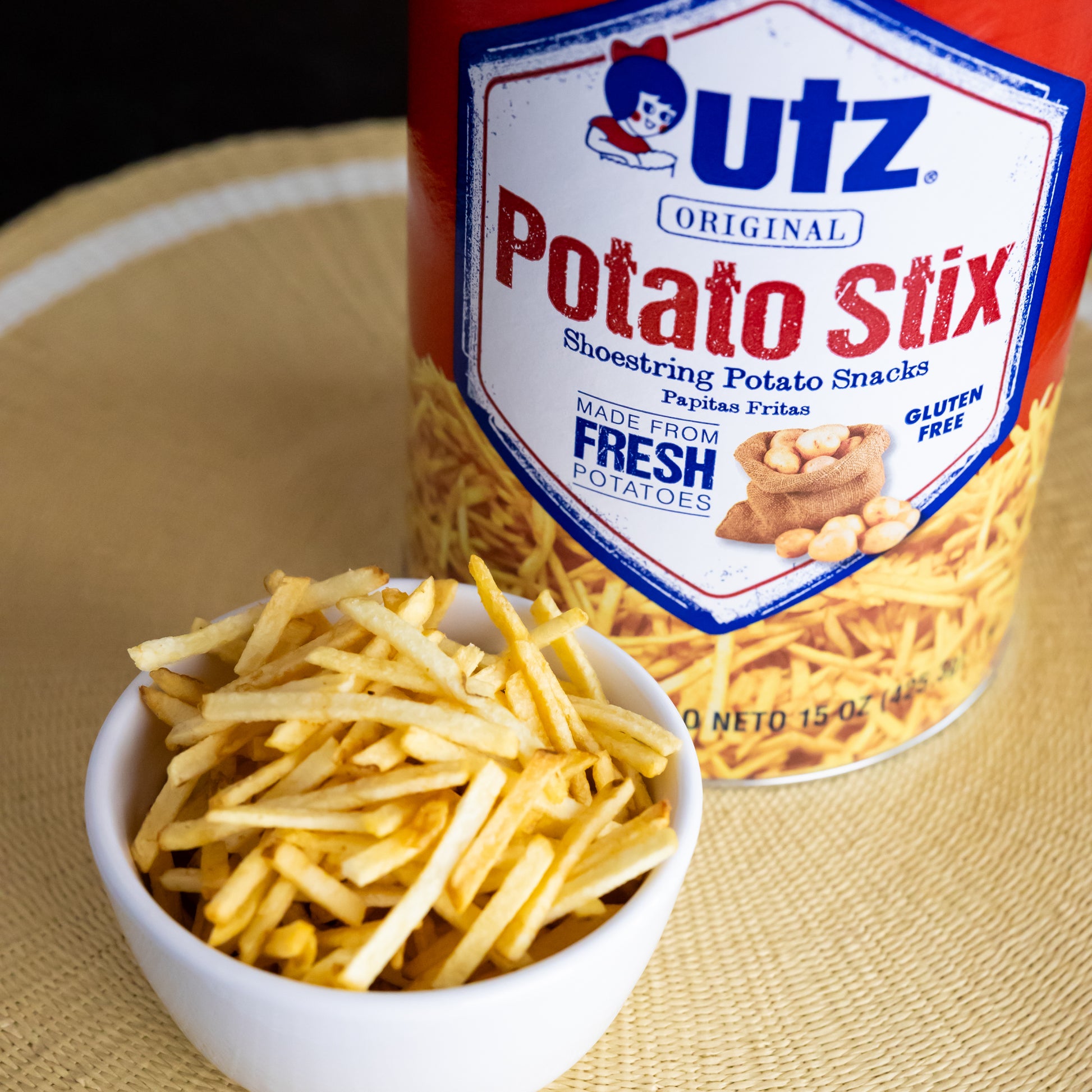 Image of a 15 oz canister of Utz Original Potato Stix next to a small white bowl filled with the shoestring potato sticks. The label on the canister reads 'Made from Fresh Potatoes' and 'Gluten Free.'