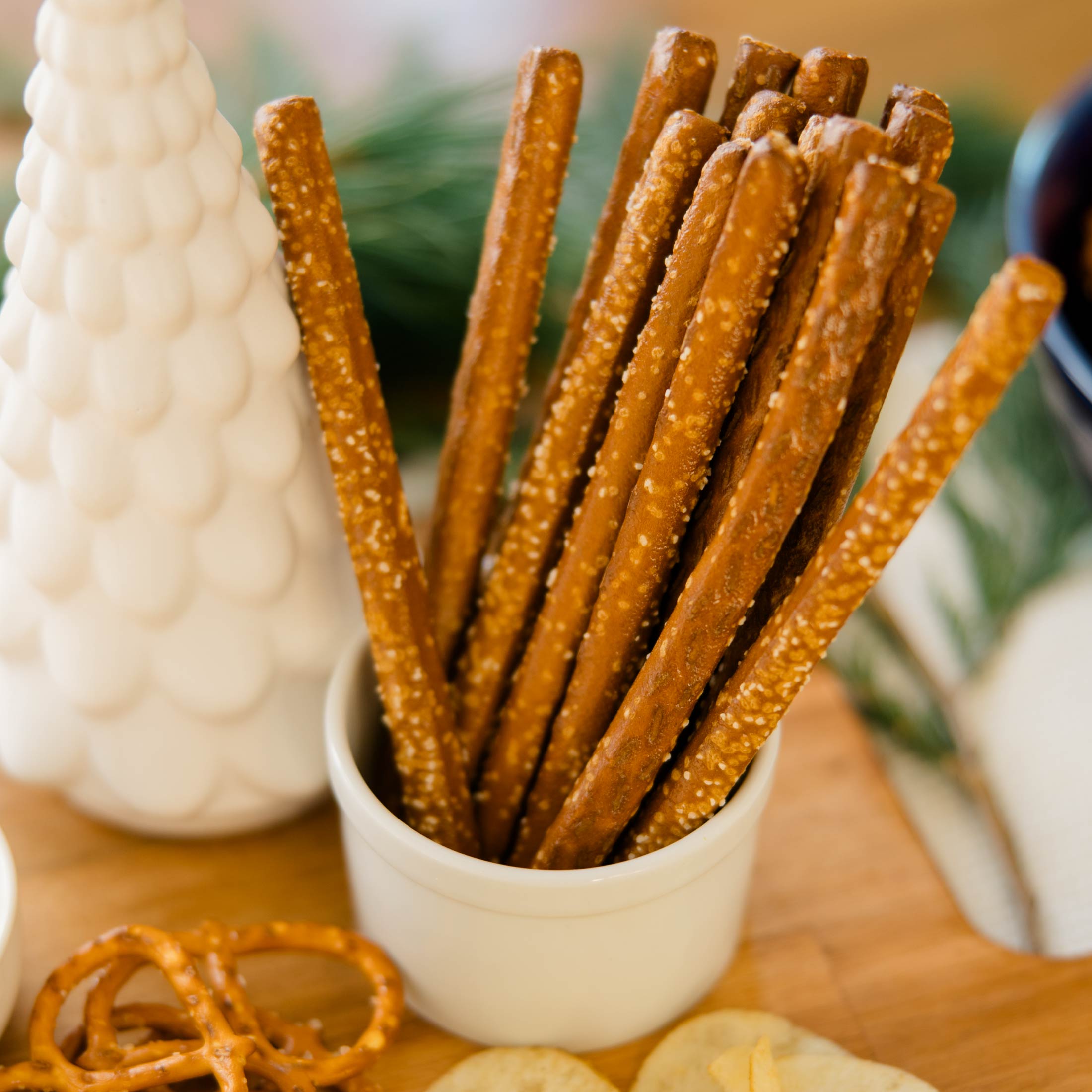 Pretzel sticks in a white container on a wooden surface with decorative items in the background.