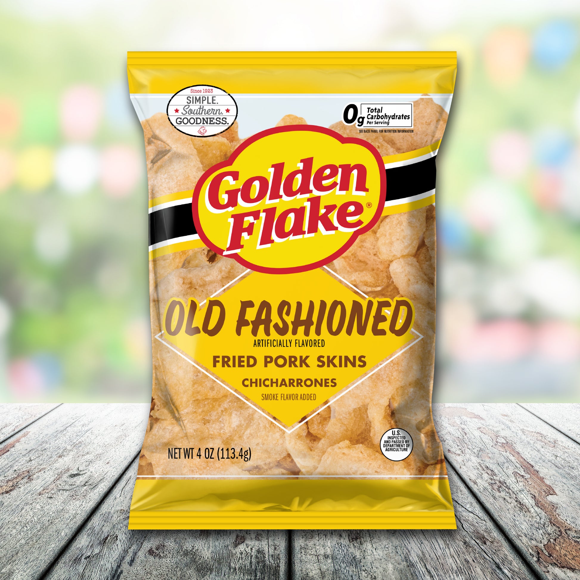 Packaging of Golden Flake Old Fashioned Fried Pork Skins on a wooden surface with a blurred colorful background.