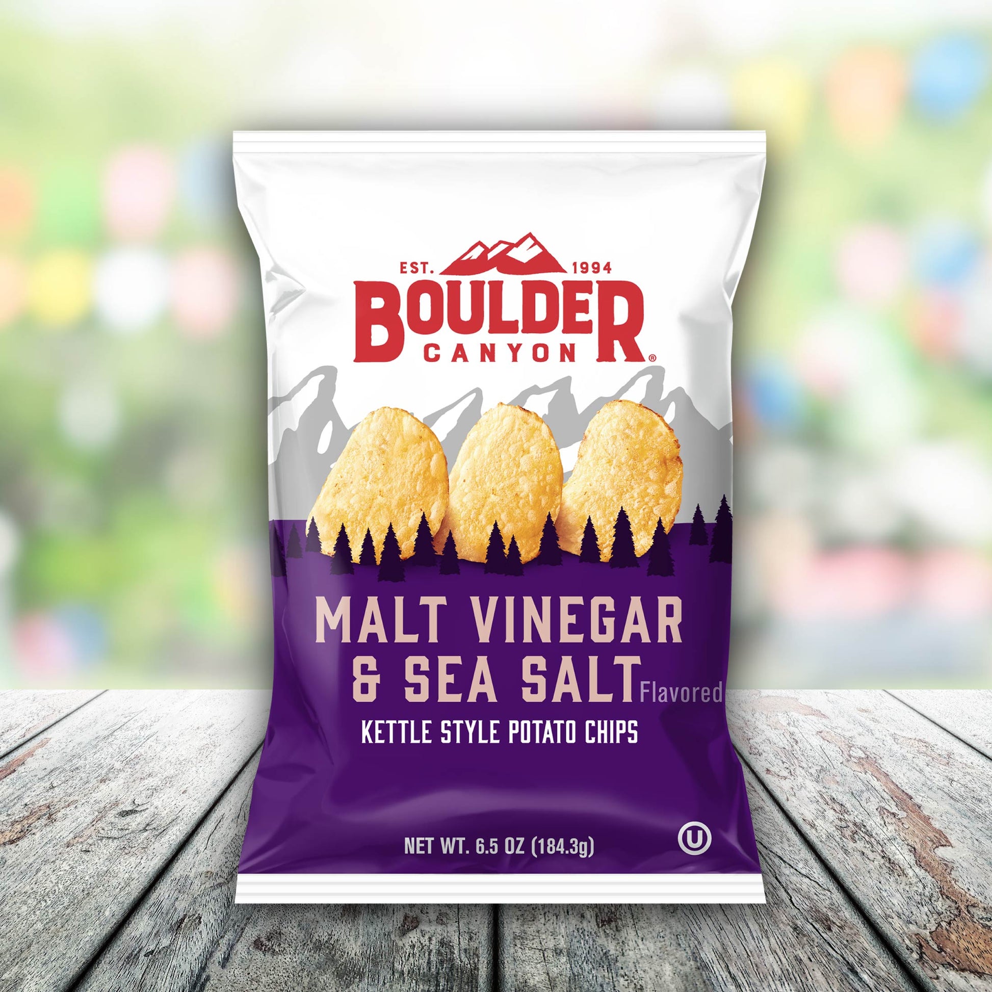 Boulder Canyon Malt Vinegar & Sea Salt kettle style potato chips packaging on a wooden surface with a blurred colorful background.
