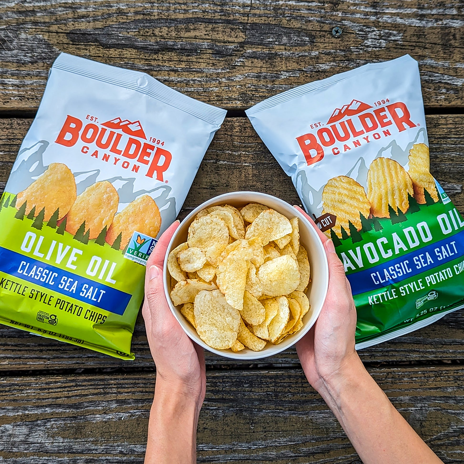 Two bags of Boulder Canyon potato chips and a bowl of chips on a wooden surface.