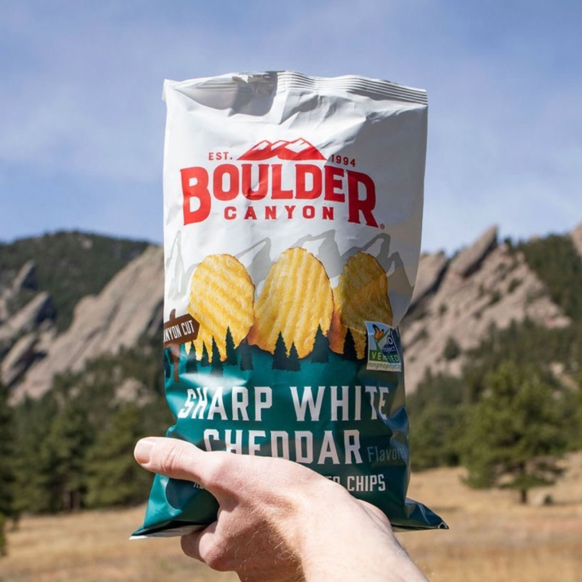 Hand holding a Boulder Canyon Sharp White Cheddar chip bag with mountains in the background