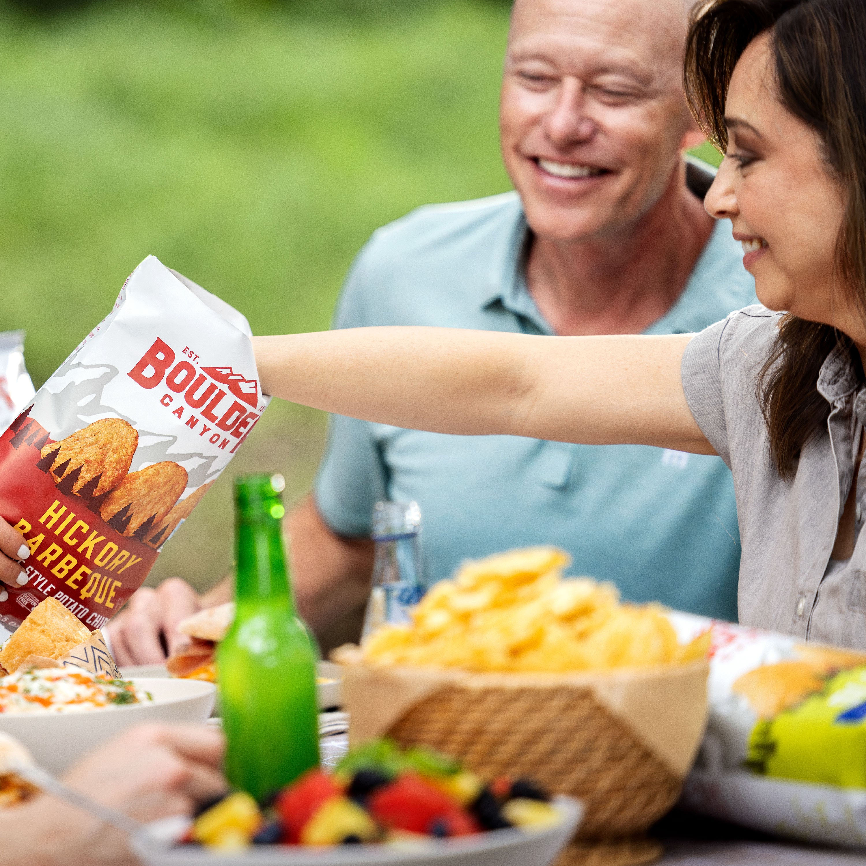 Woman reaching for Boulder Canyon Hickory Barbecue chips at outdoor meal.