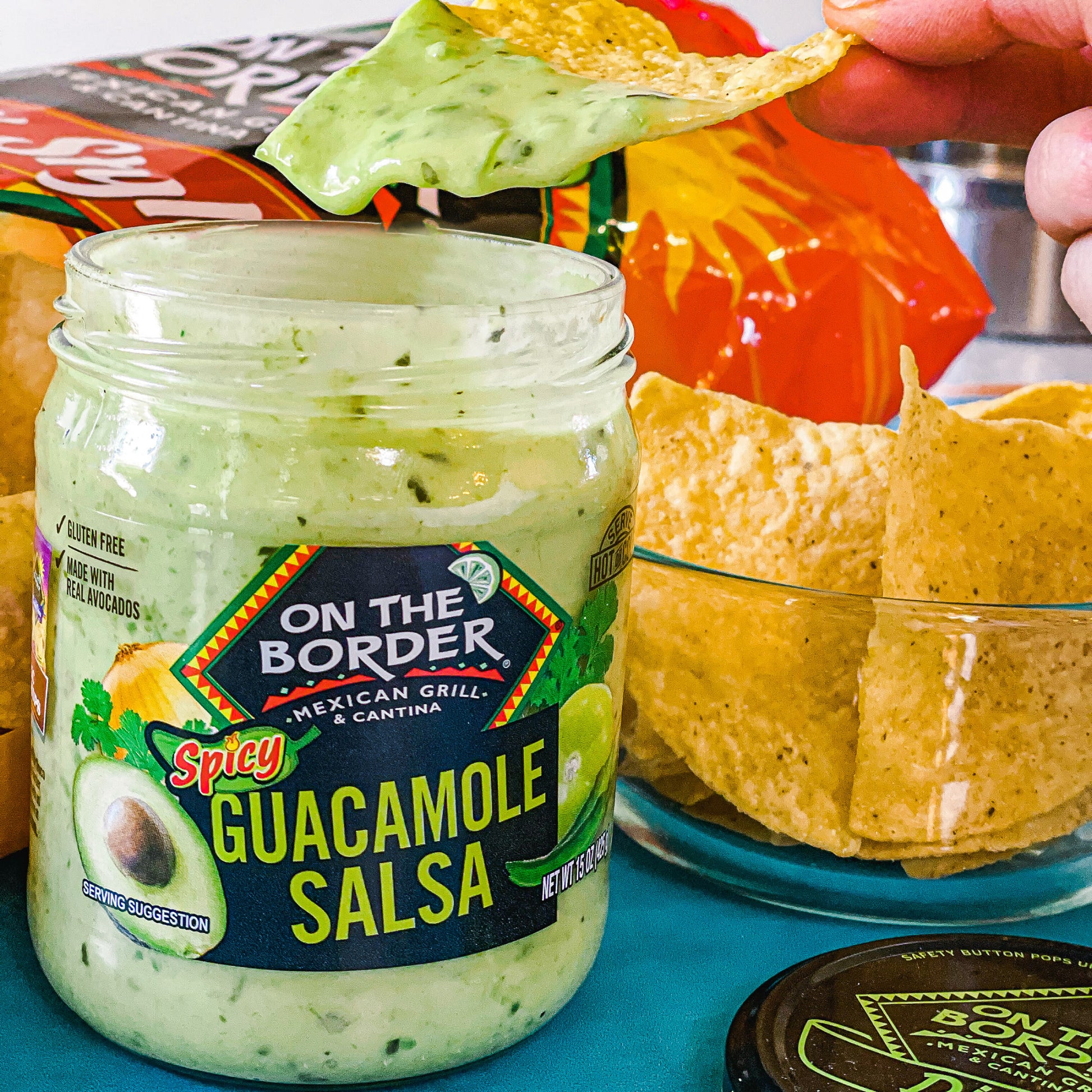 Jar of 'On The Border' Spicy Guacamole Salsa with a tortilla chip being dipped into it, surrounded by chips and salsa containers.