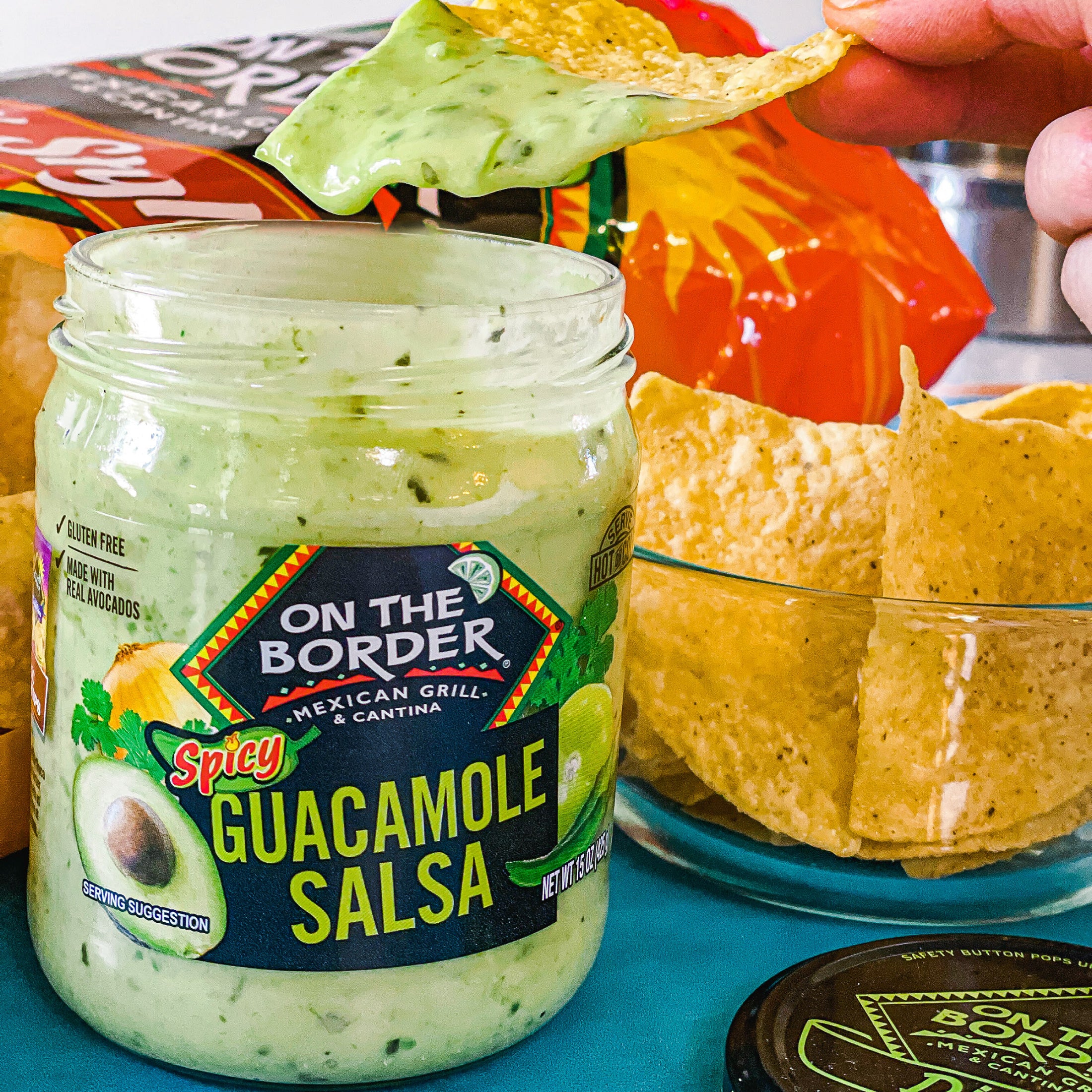 Jar of 'On The Border' Spicy Guacamole Salsa with a tortilla chip being dipped into it, surrounded by chips and salsa containers.