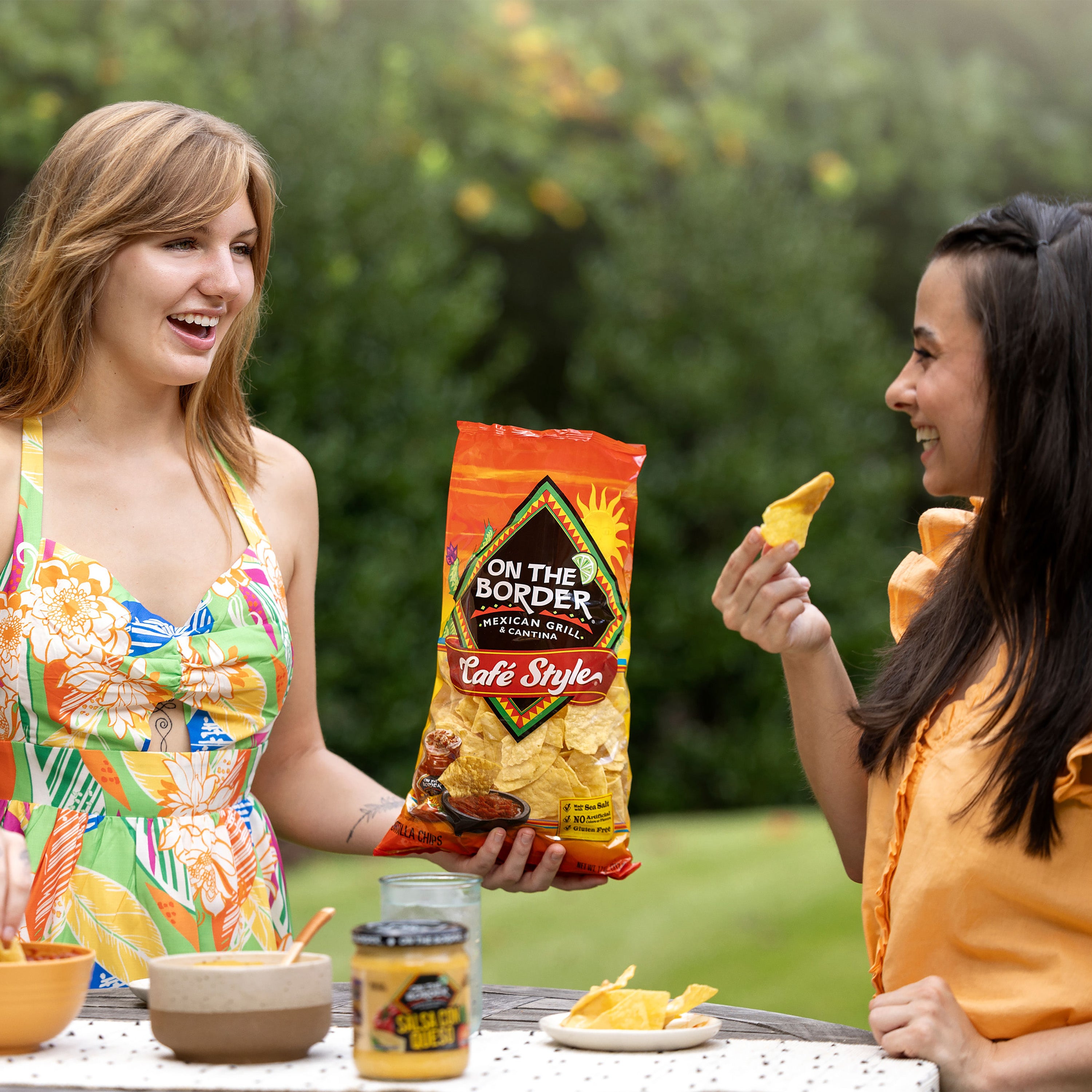 Two women enjoying chips from 'On The Border' at a picnic table outdoors.