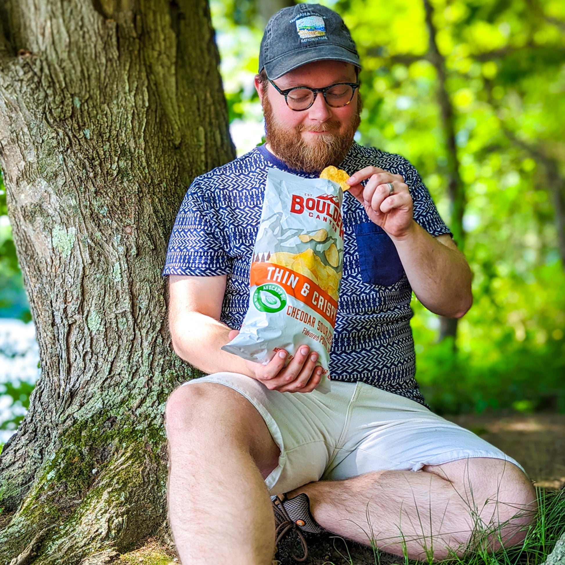 Man sitting under a tree in a forest, eating snacks from a Boulder Canyon bag.
