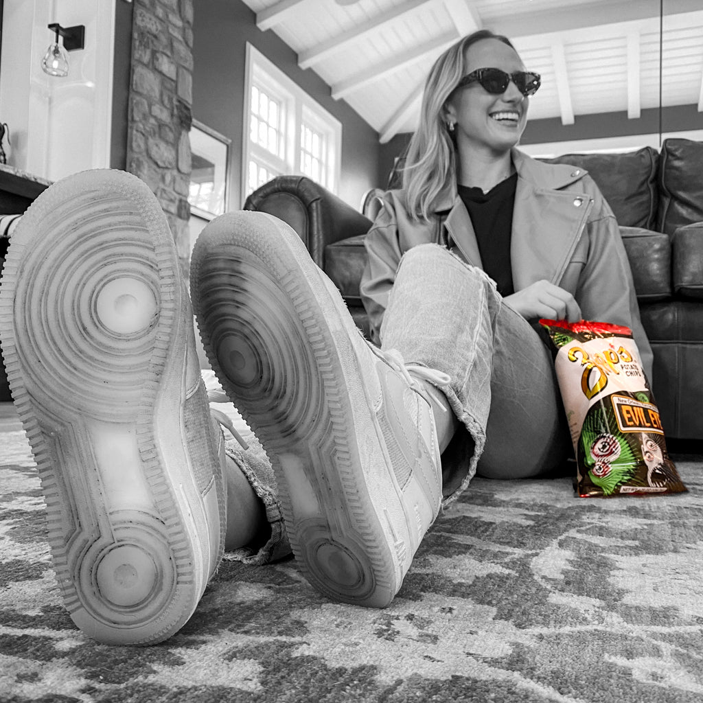 Person wearing large sneakers sitting on a carpeted floor with a bag of chips nearby.