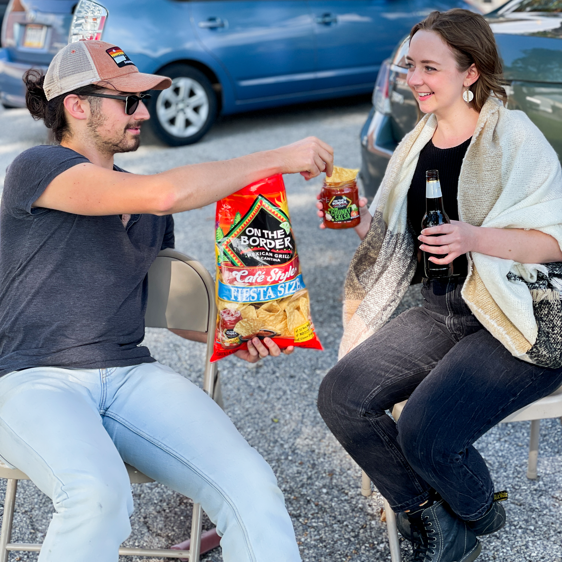 Two people sitting outdoors, one with a bag of 'On the Border' chips and a bottle, the other with a jar of salsa.