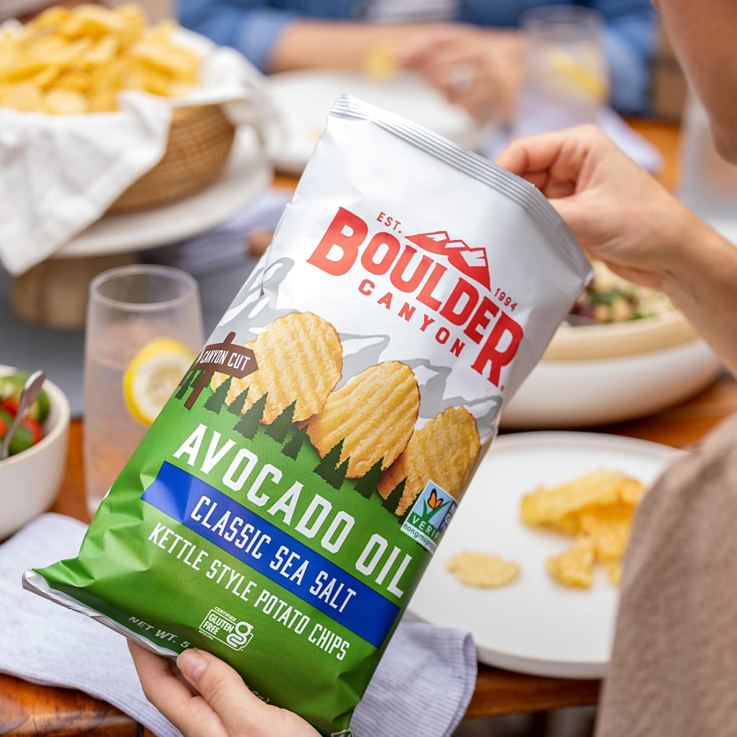 Person holding a bag of Boulder Canyon avocado oil kettle-style potato chips at a casual dining setting.