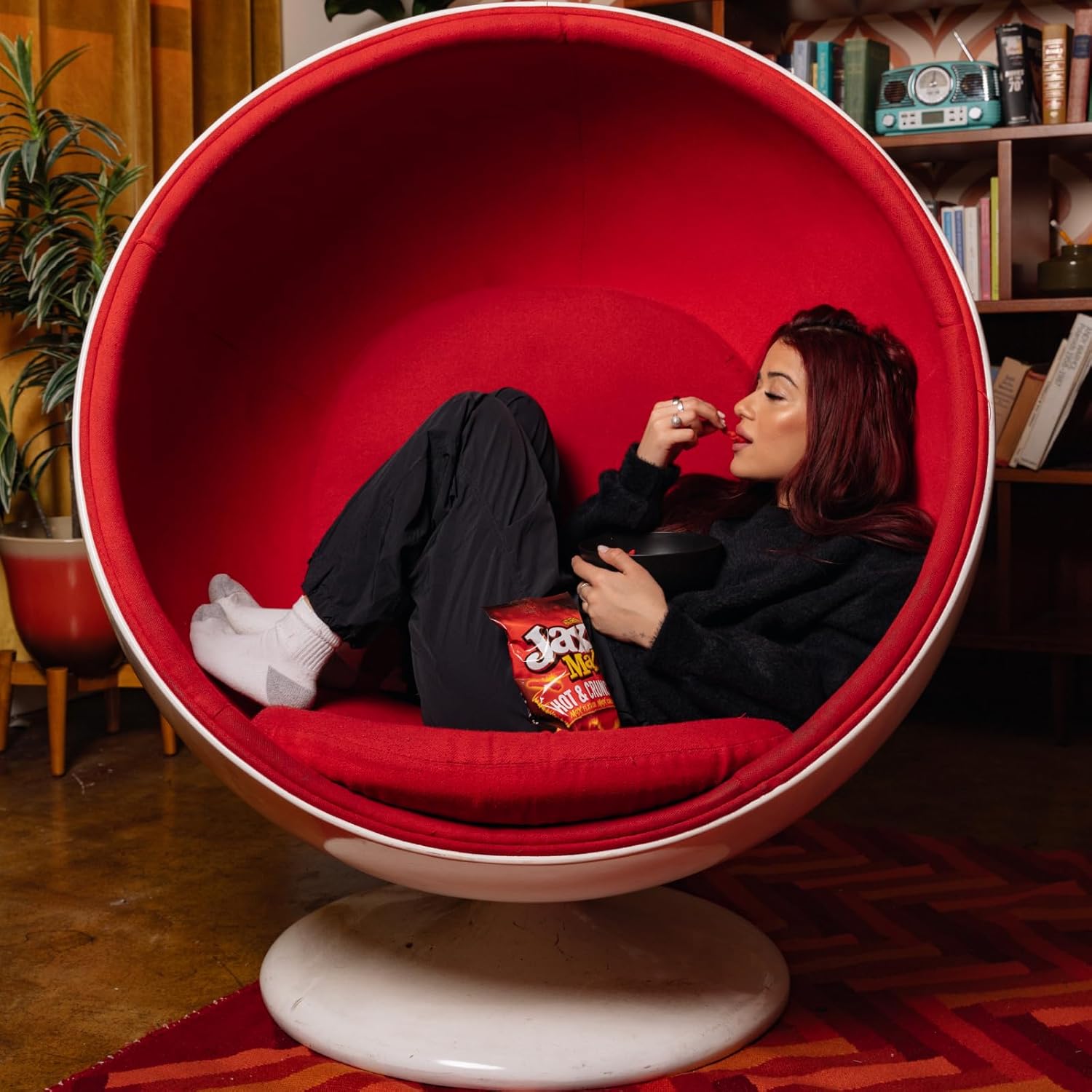 Person sitting in a red egg chair holding a bag of chips in a cozy room.