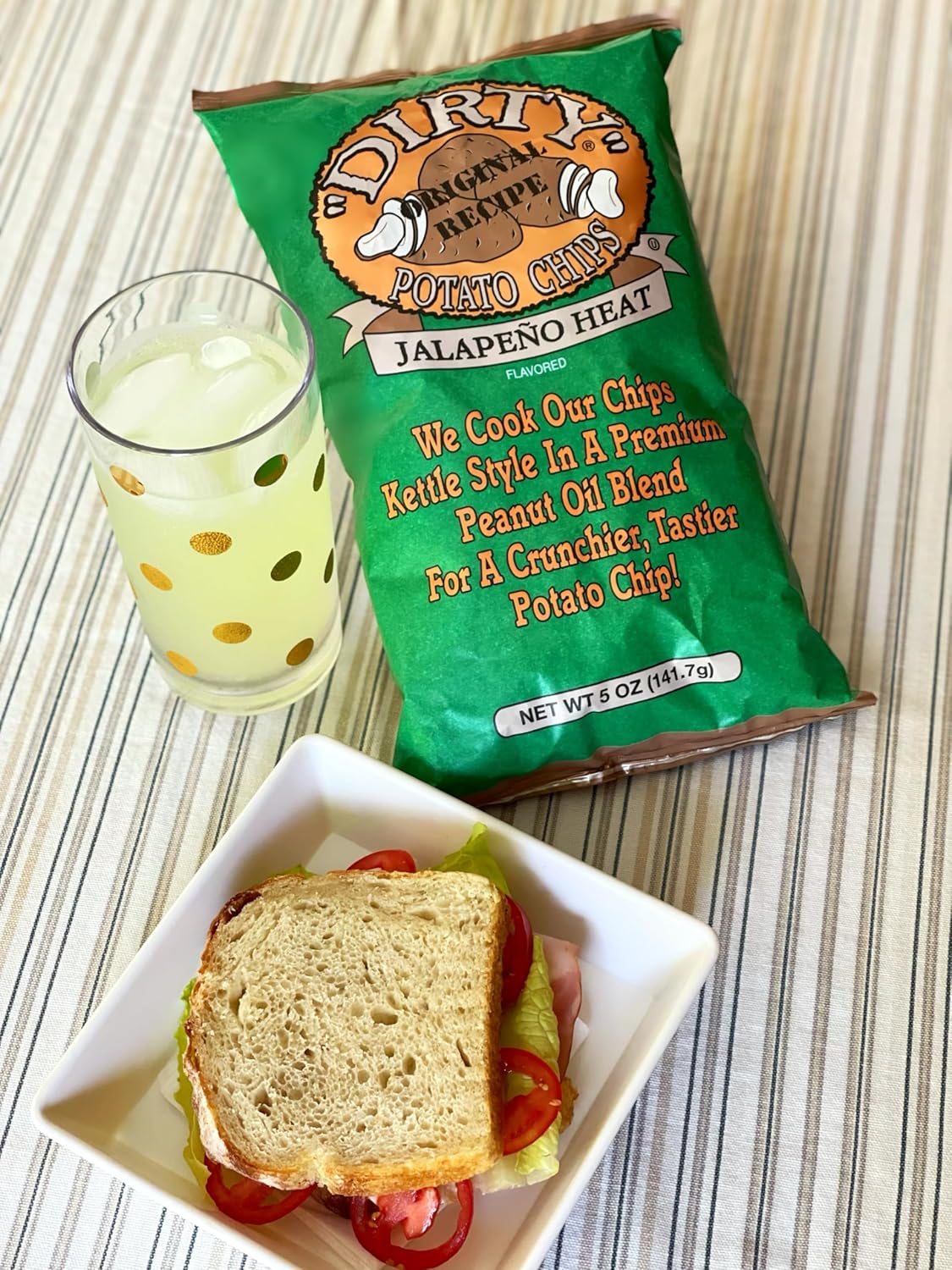 Green bag of Dirty Potato Chips with a glass of lemonade and a sandwich on a striped surface
