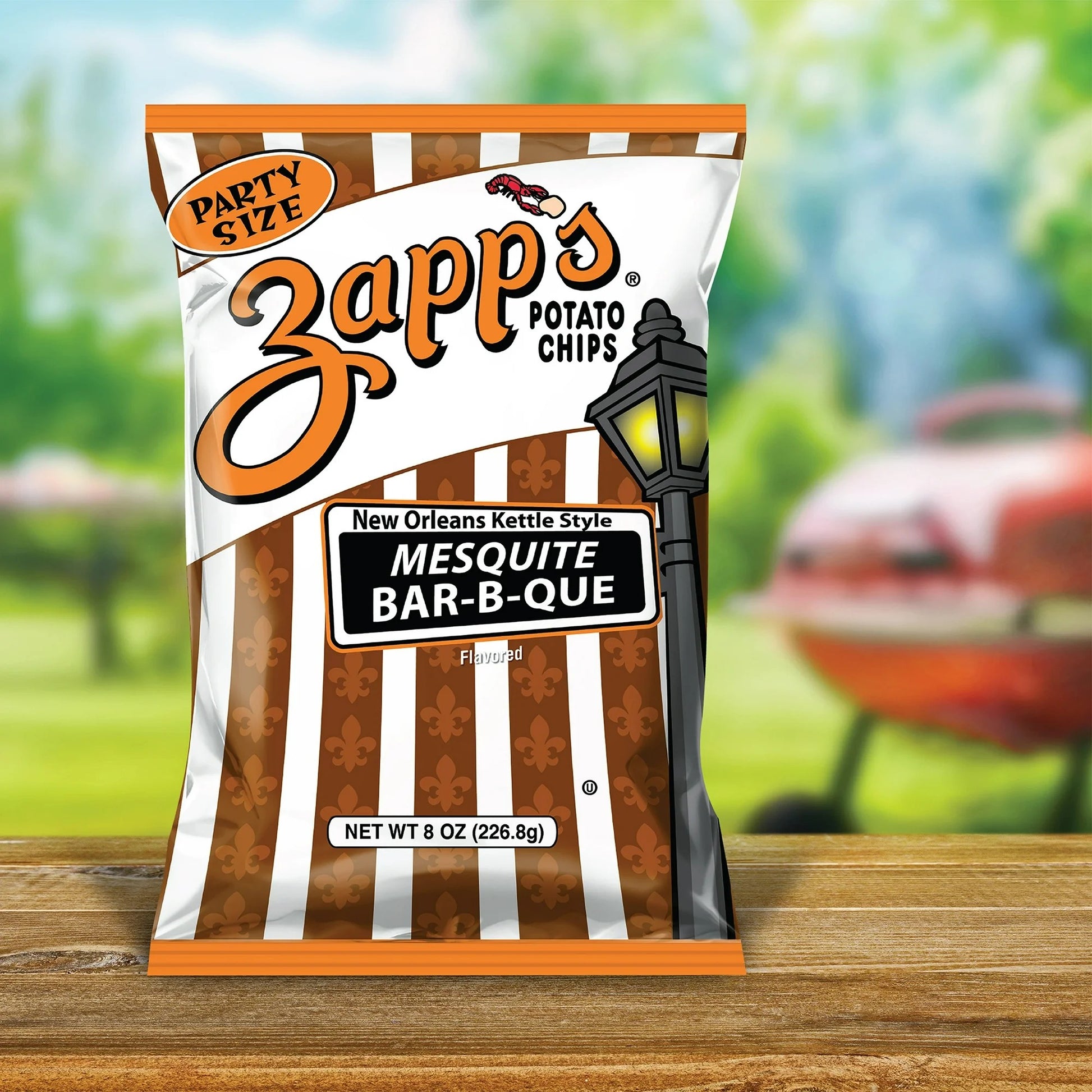 Zapp's potato chips bag with mesquite bar-b-que flavor on a wooden surface with a blurred grill and grass background.