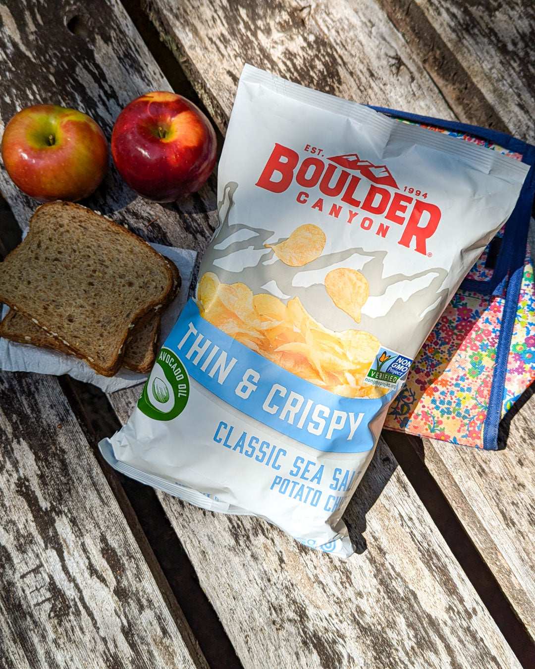 Boulder Canyon thin and crispy potato chips on a wooden surface with apples and toast.