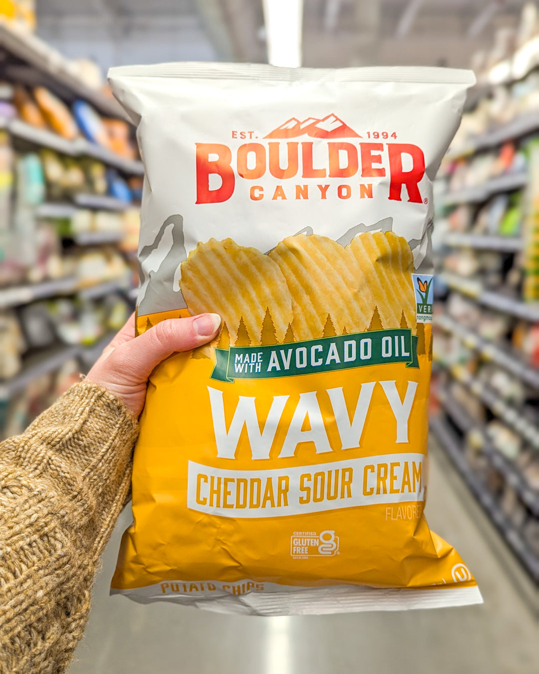 Bag of Boulder Canyon Wavy Cheddar Sour Cream chips held in a grocery store.