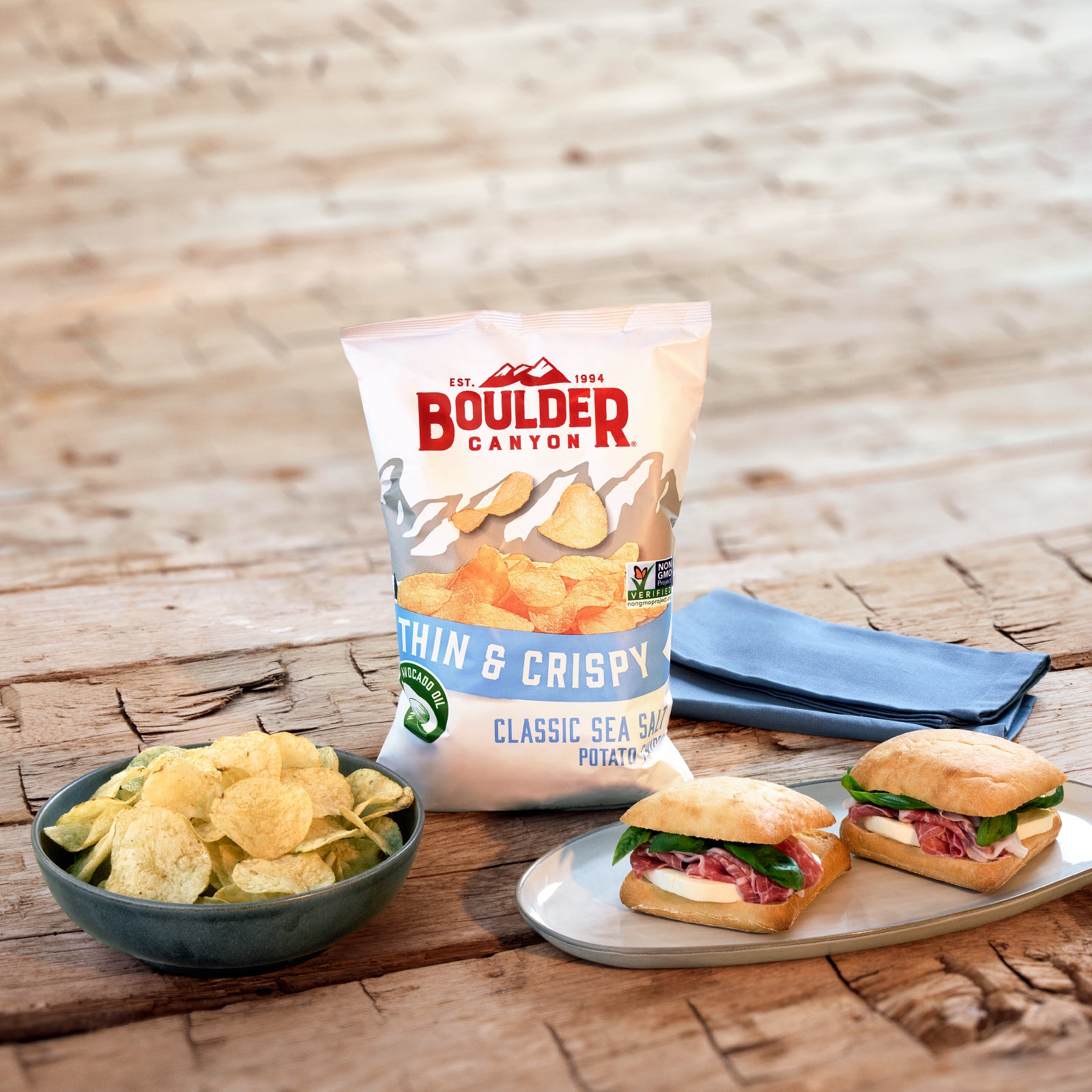 Boulder Canyon thin crispy potato chips with a bowl of chips and sandwiches on a wooden surface