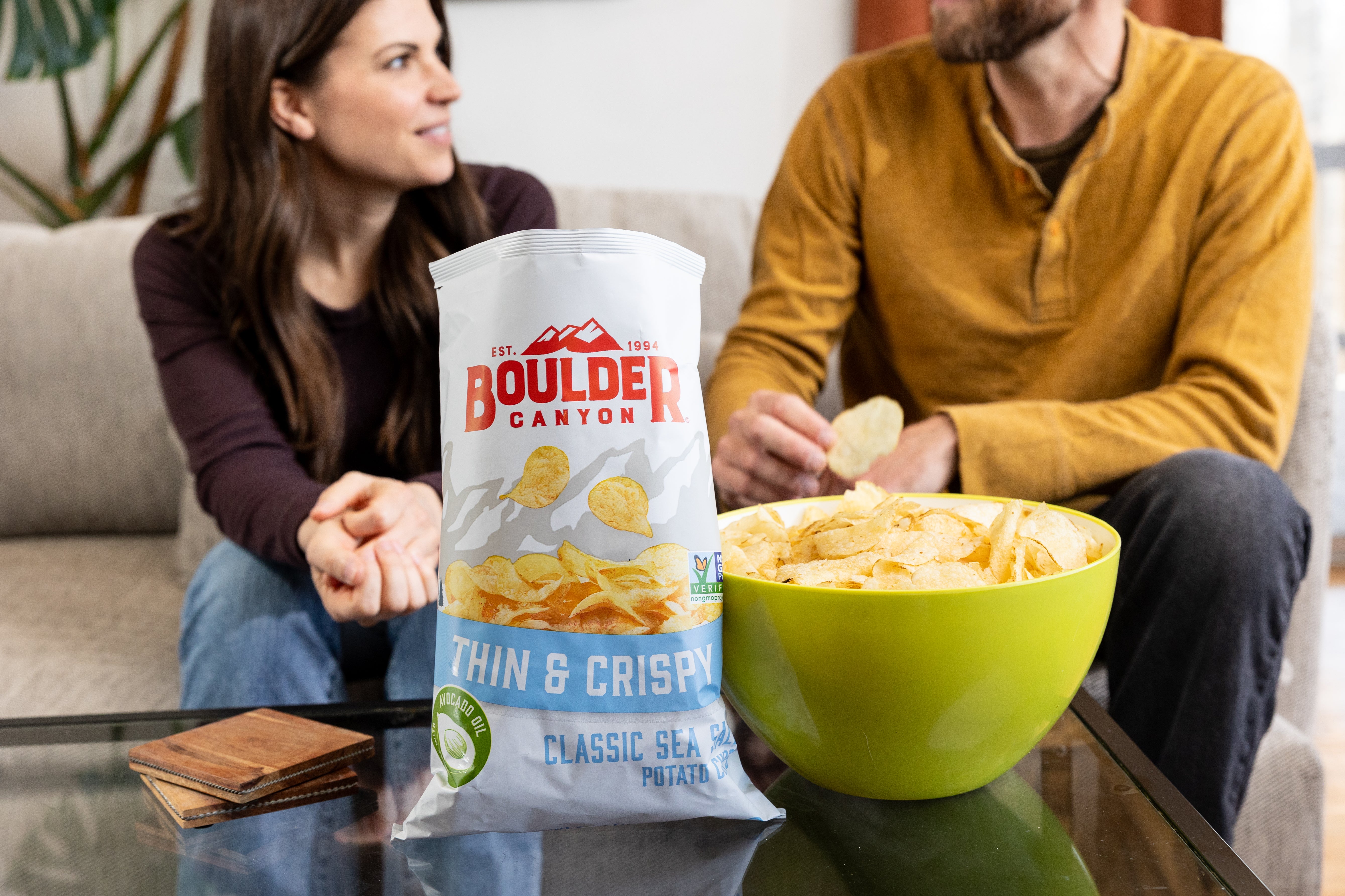 Two people sitting on a couch with a bag of Boulders Canyon thin and crispy chips and a bowl of chips on a table.
