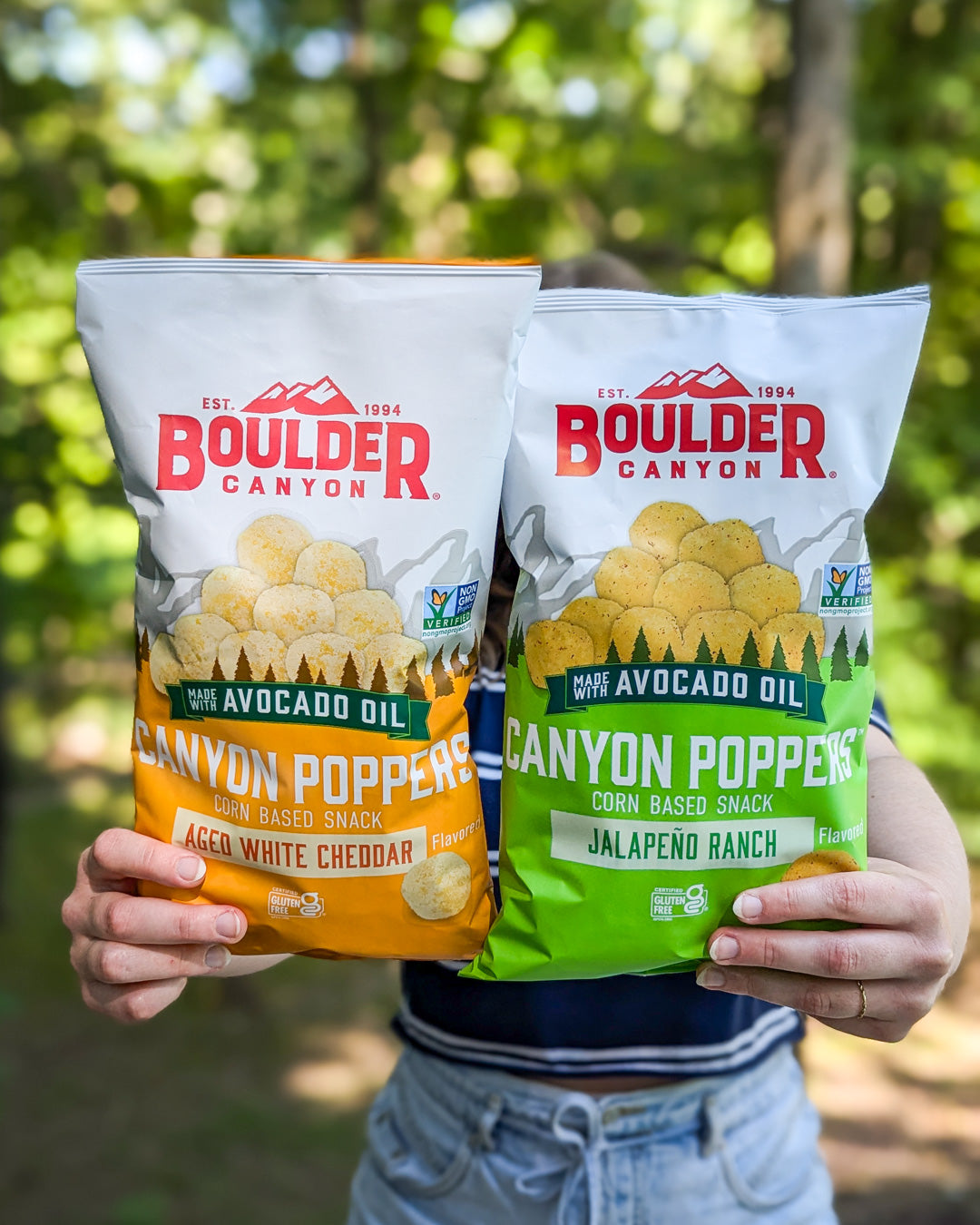 Two bags of Boulder Canyon Canyon Poppers snack held against a blurred natural background