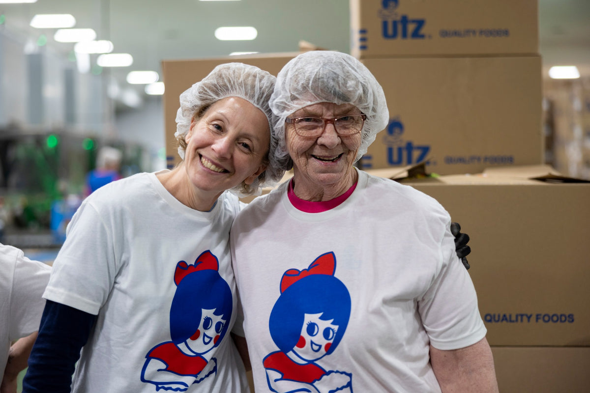 Two smiling Utz team members in hairnets and Little Utz Girl t-shirts on the production floor, with Utz Quality Foods cases stacked behind them