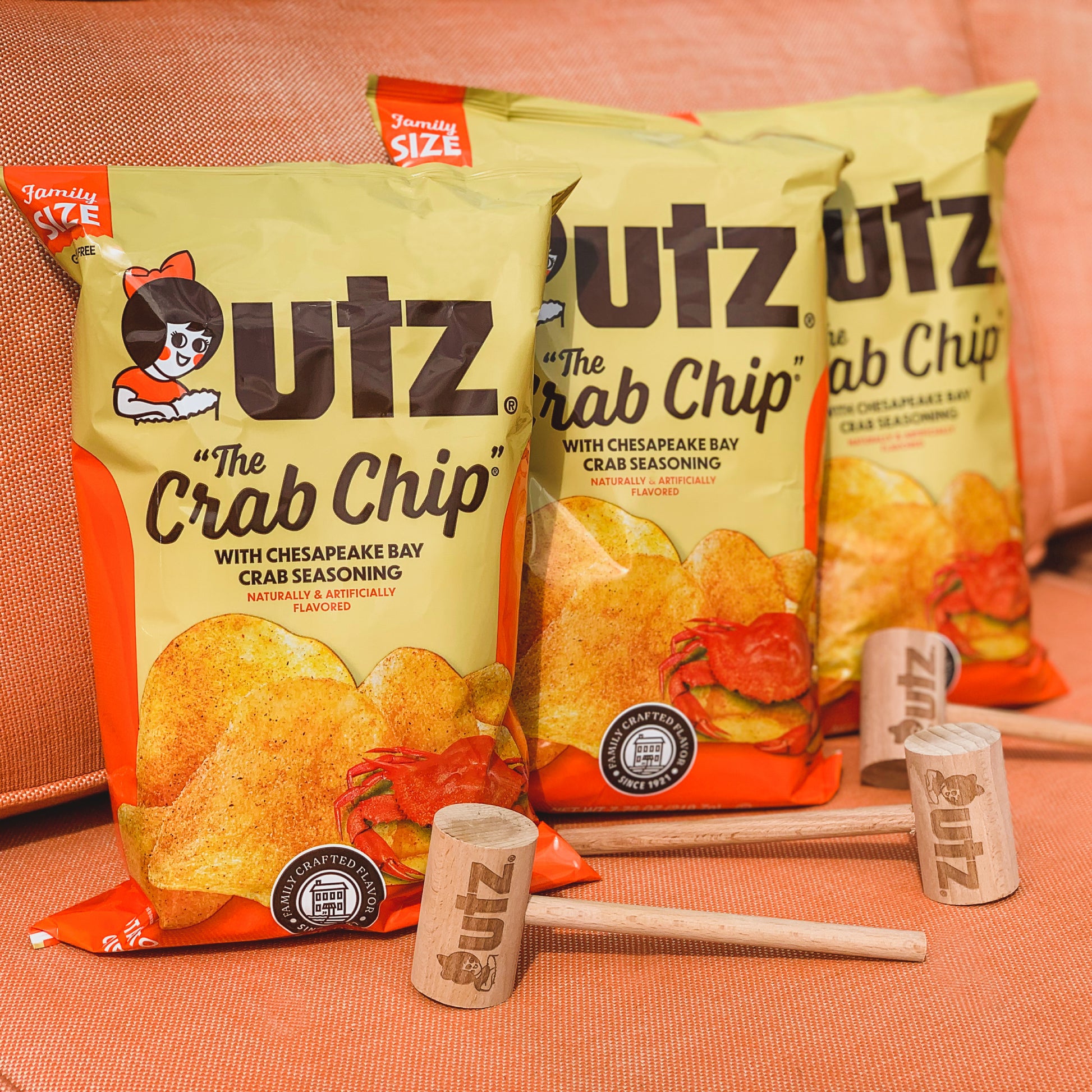 Three bags of Utz 'The Crab Chip' with a hammer and can opener on an orange surface.