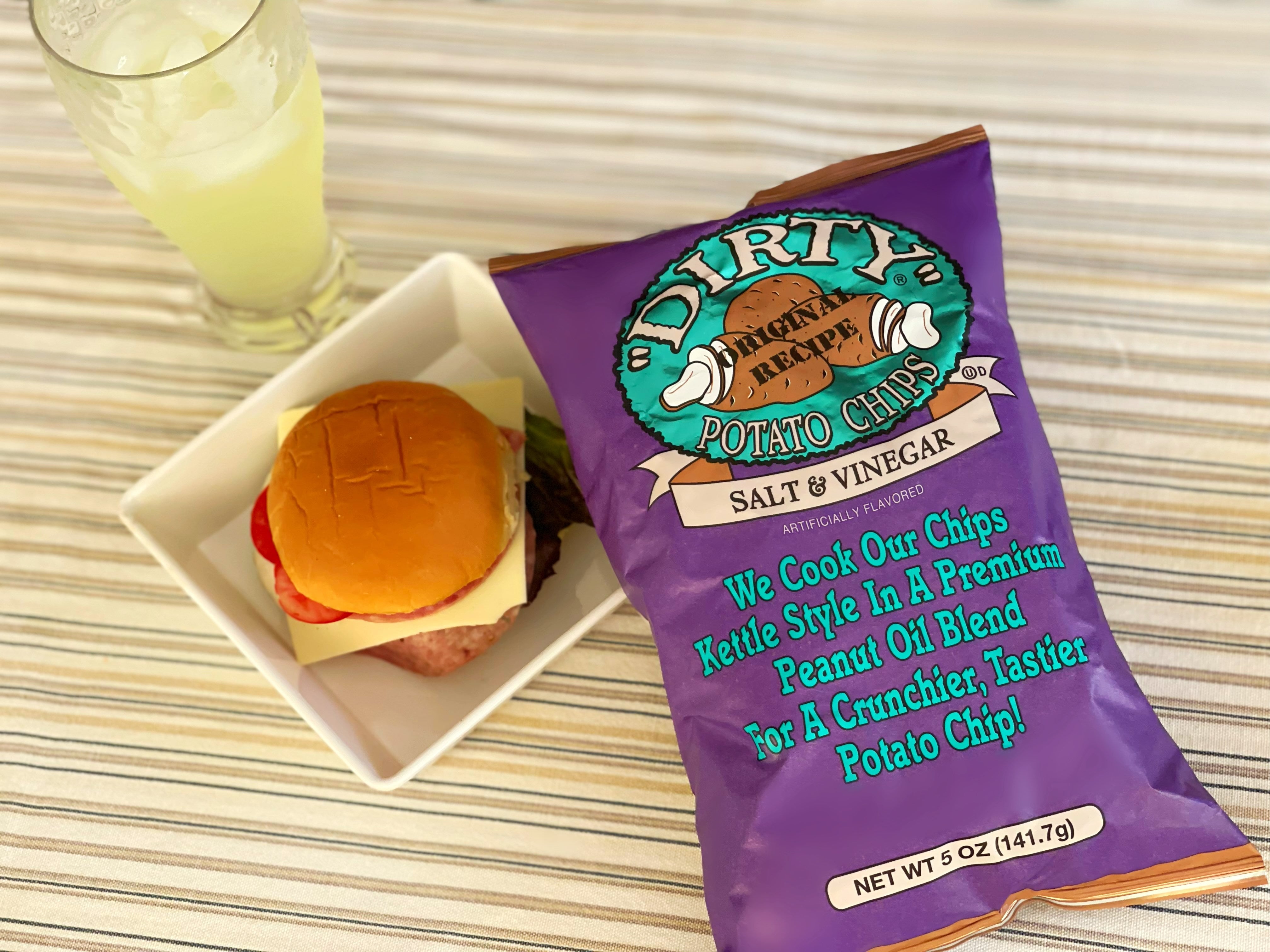 Purple bag of Dirty Potato Chips next to a small plate with a sandwich on a striped tablecloth.