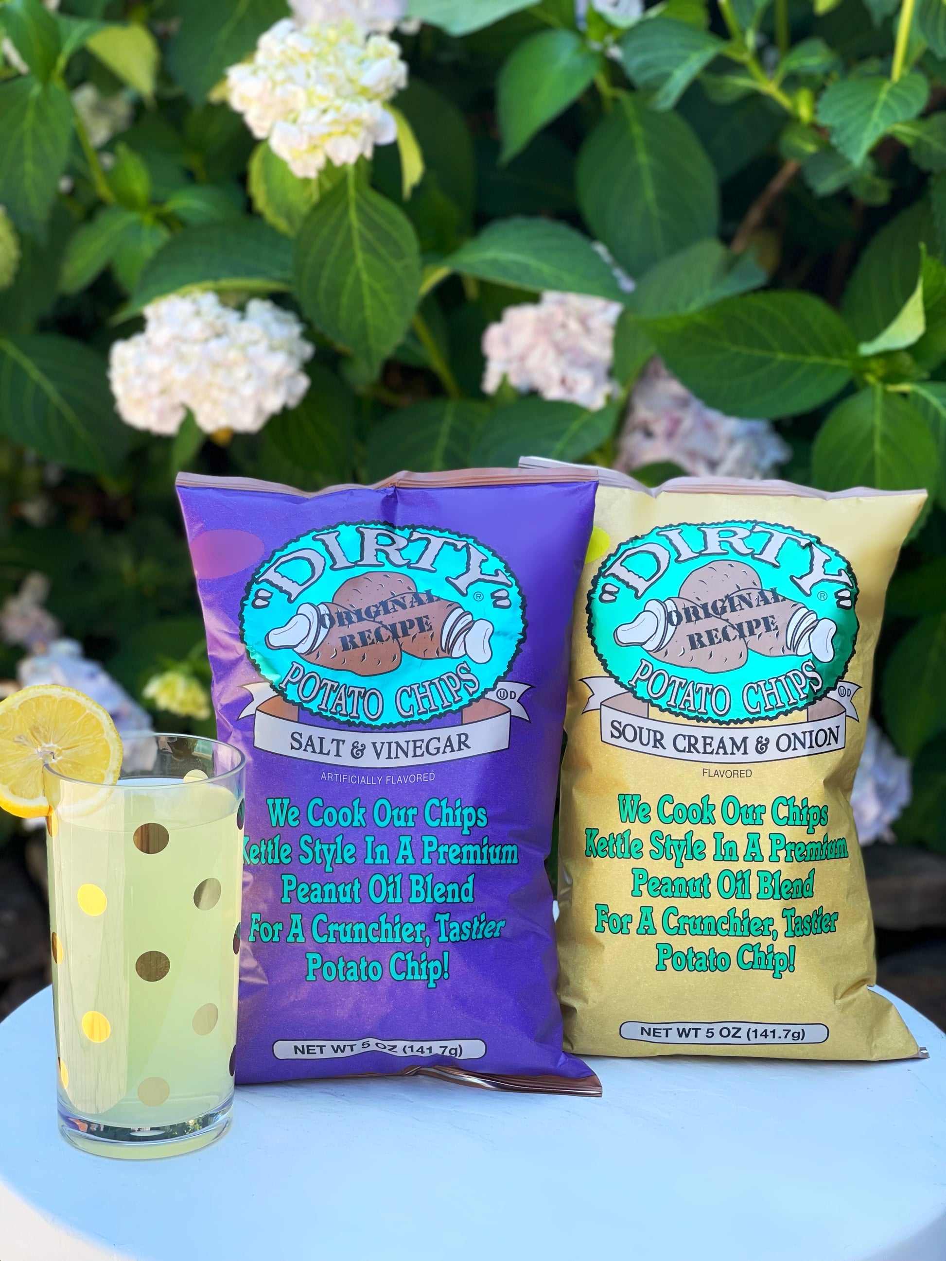 Two bags of Dirty Original Recipe potato chips with a drink on a table outdoors.