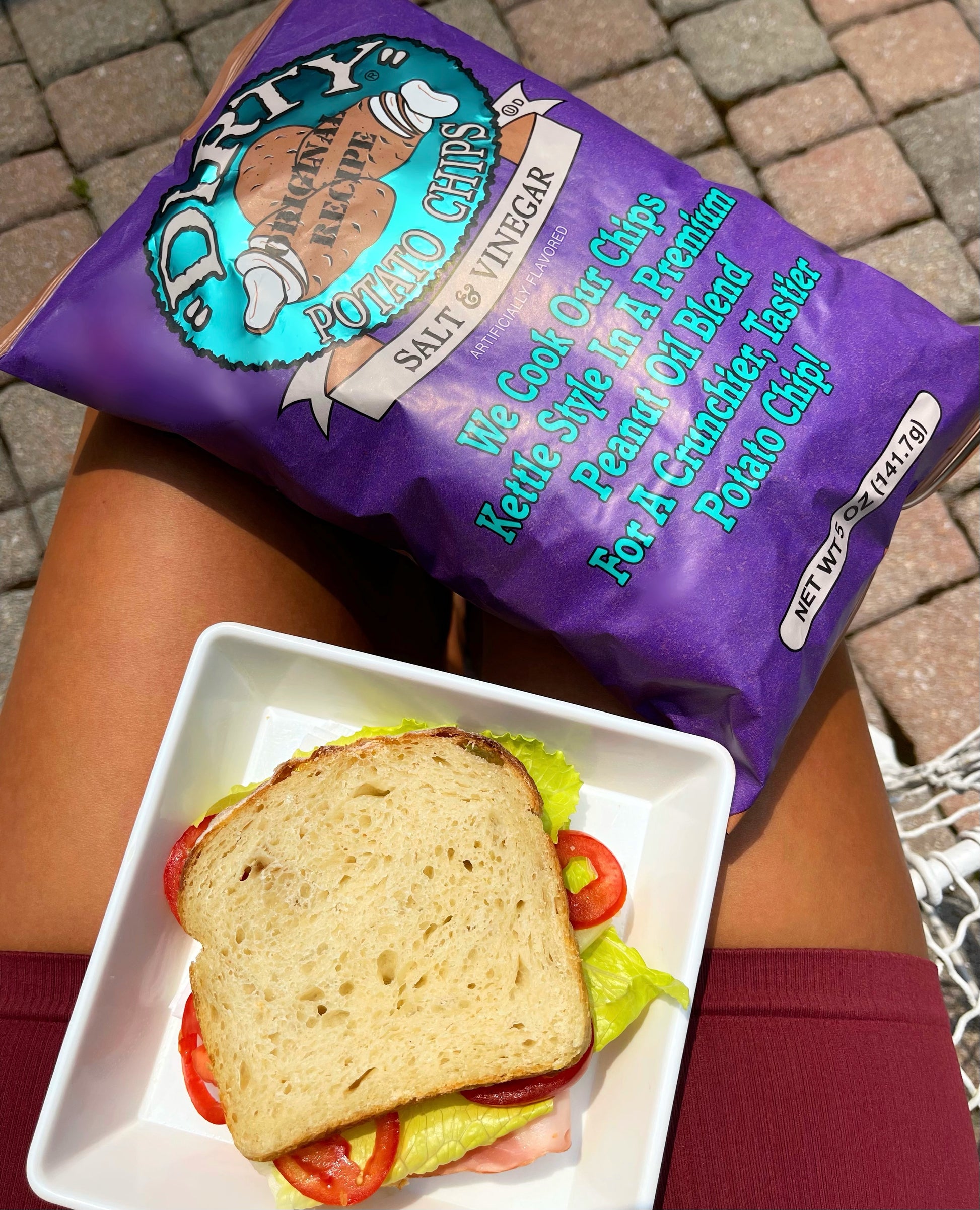 Purple bag of 'Dirty Chips' potato chips next to a sandwich on a white plate.