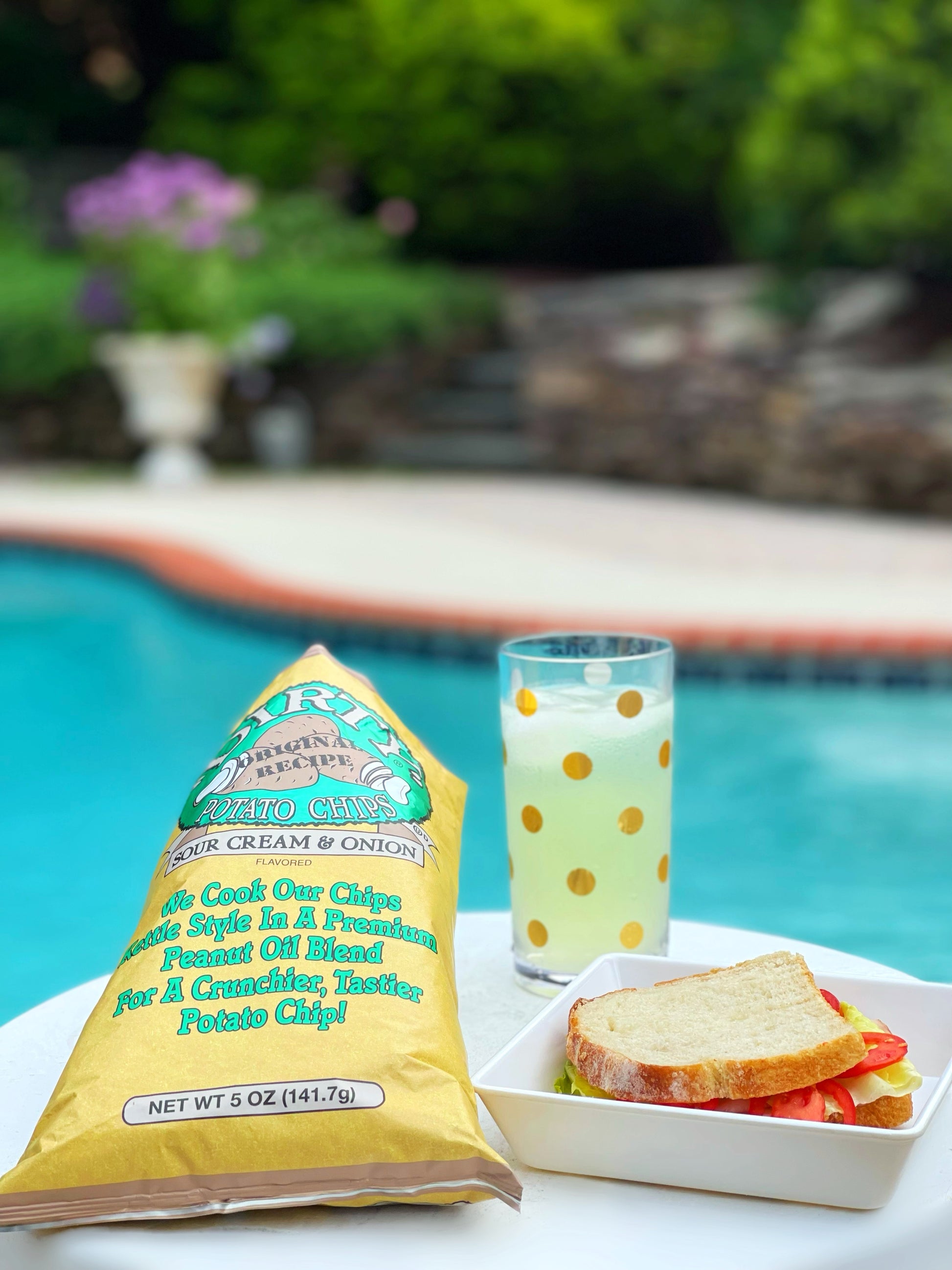 Packaged potato chips, a glass with a polka dot pattern, and a sandwich on a poolside table.