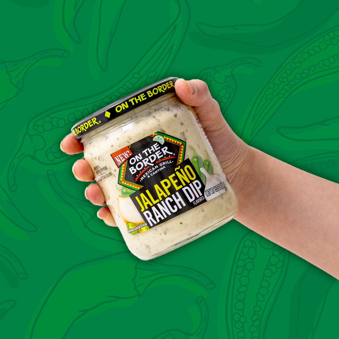 Hand holding a container of 'On The Border' Jalapeno Ranch Dip against a green background