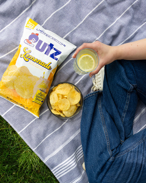 Person sitting on a blanket with Outz Lemonade chips, a glass of lemonade, and a bowl of chips.