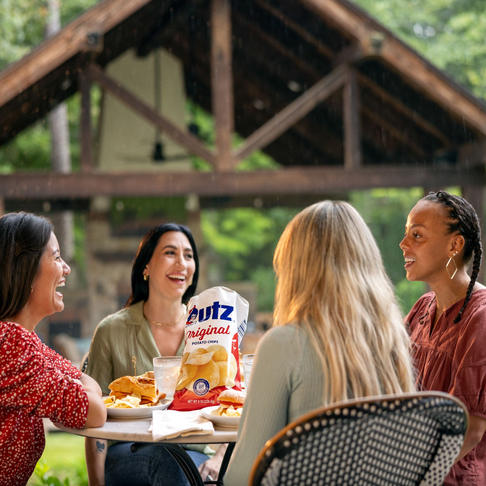 Four women sitting around a table outdoors, enjoying food and drinks with a bag of utz chips.