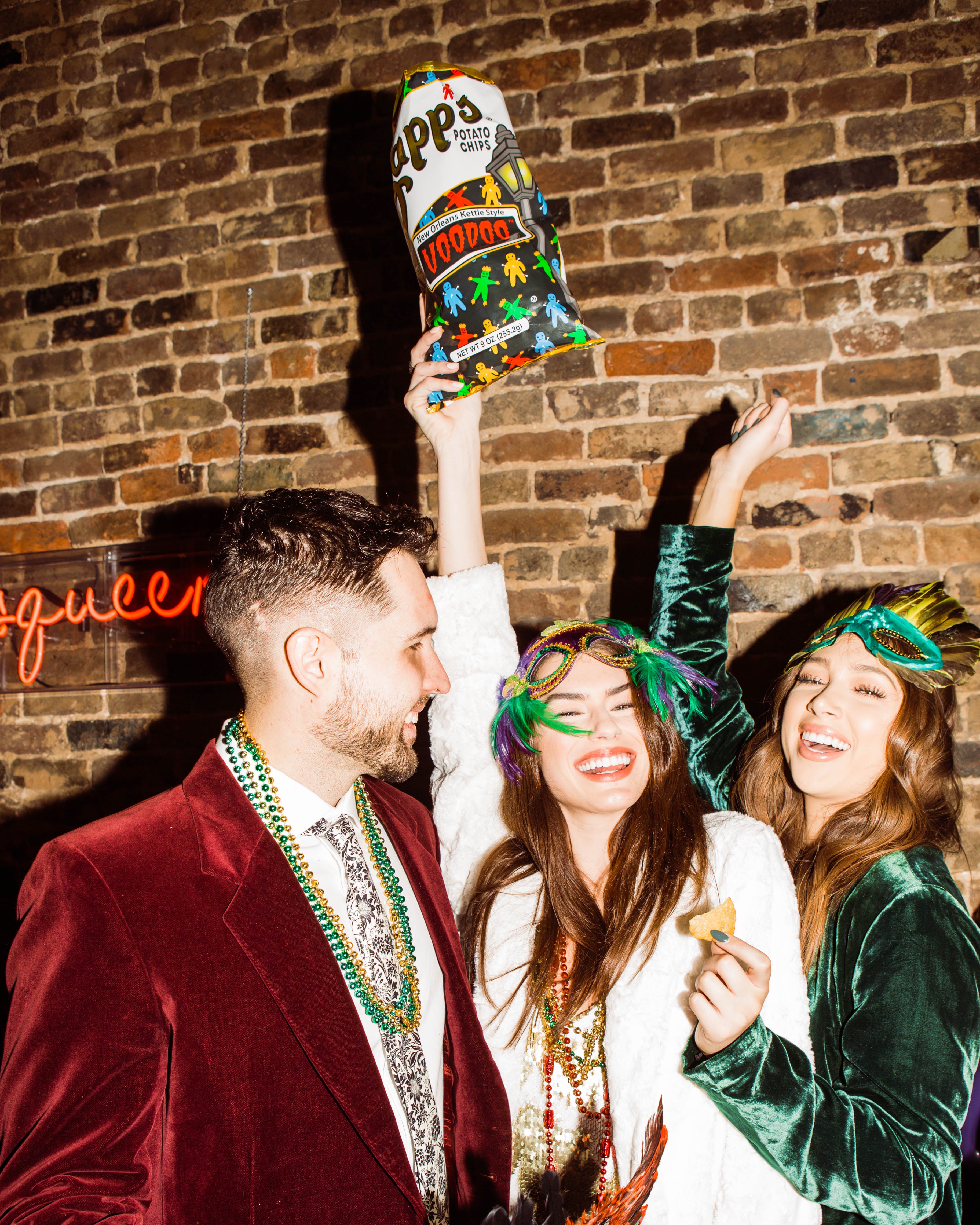 Three people at a festive event, with a woman in the center holding a bag of Zapp's Voodoo Potato Chips up in the air against a brick wall.