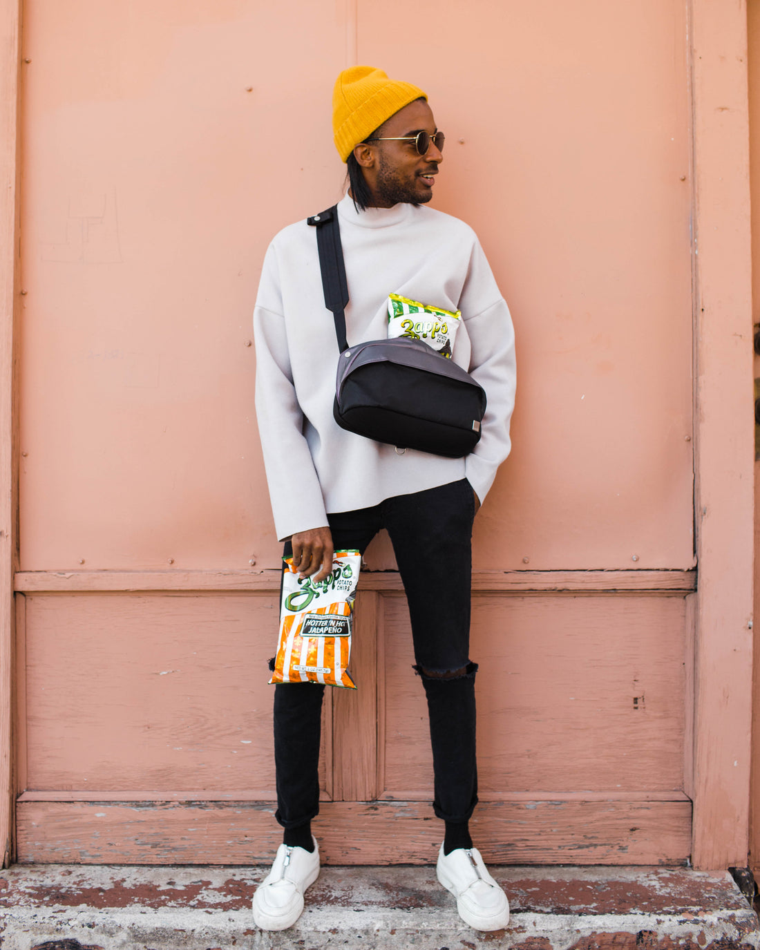 Person wearing a yellow beanie, gray sweater, black pants, and white sneakers against a pink wall.