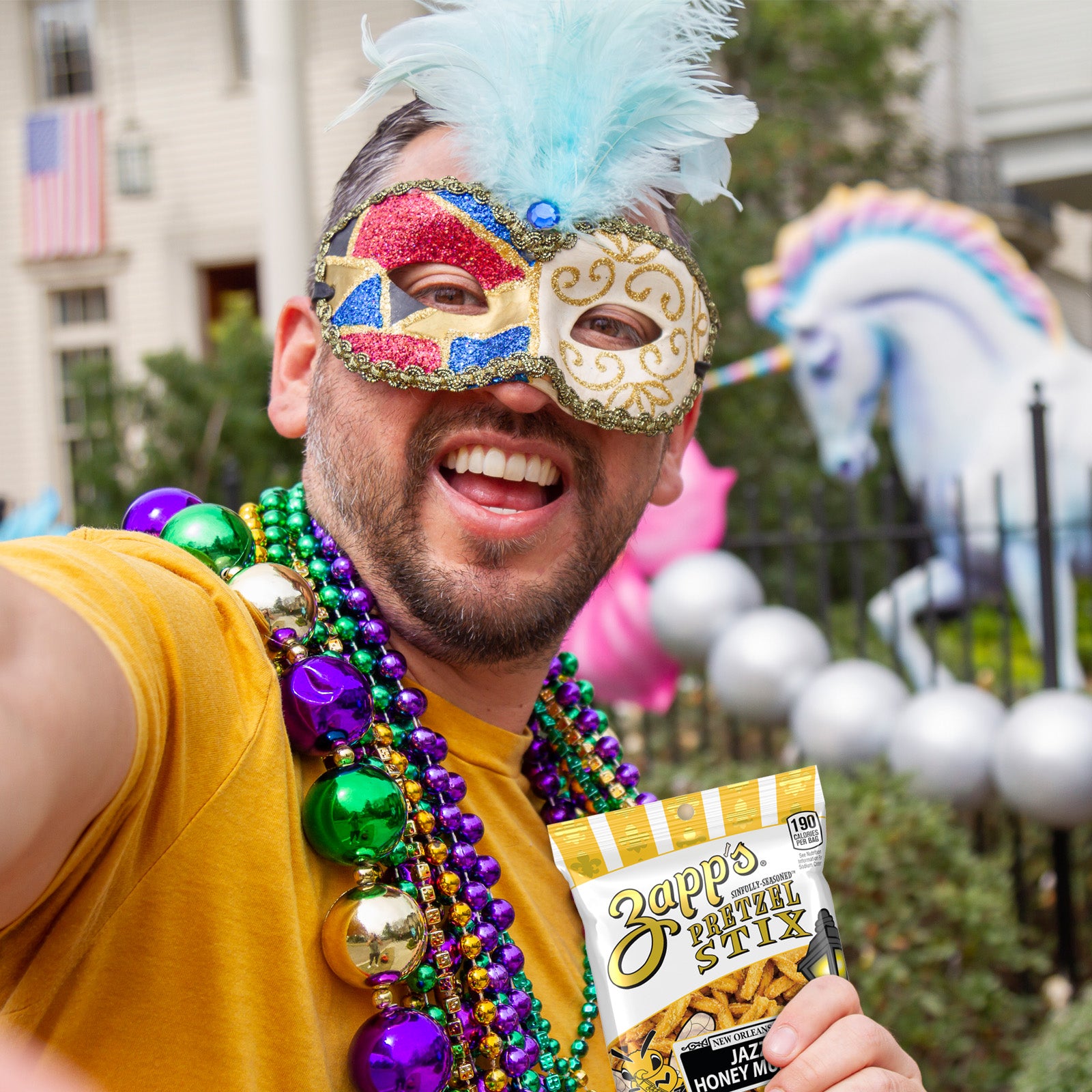 Person wearing a colorful mask and beads, holding a bag of Zappos Animal Stix, with a unicorn and balloons in the background.