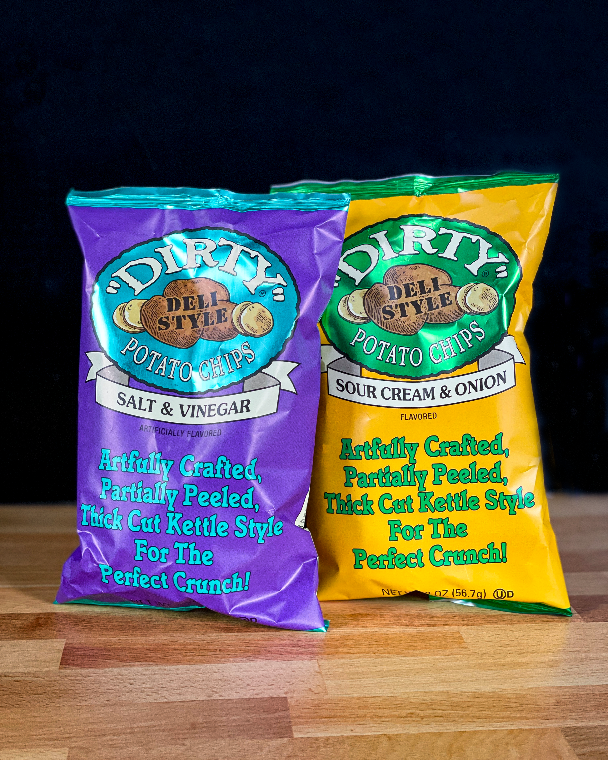 Two bags of 'Dirty' brand potato chips on a wooden surface with a dark background.