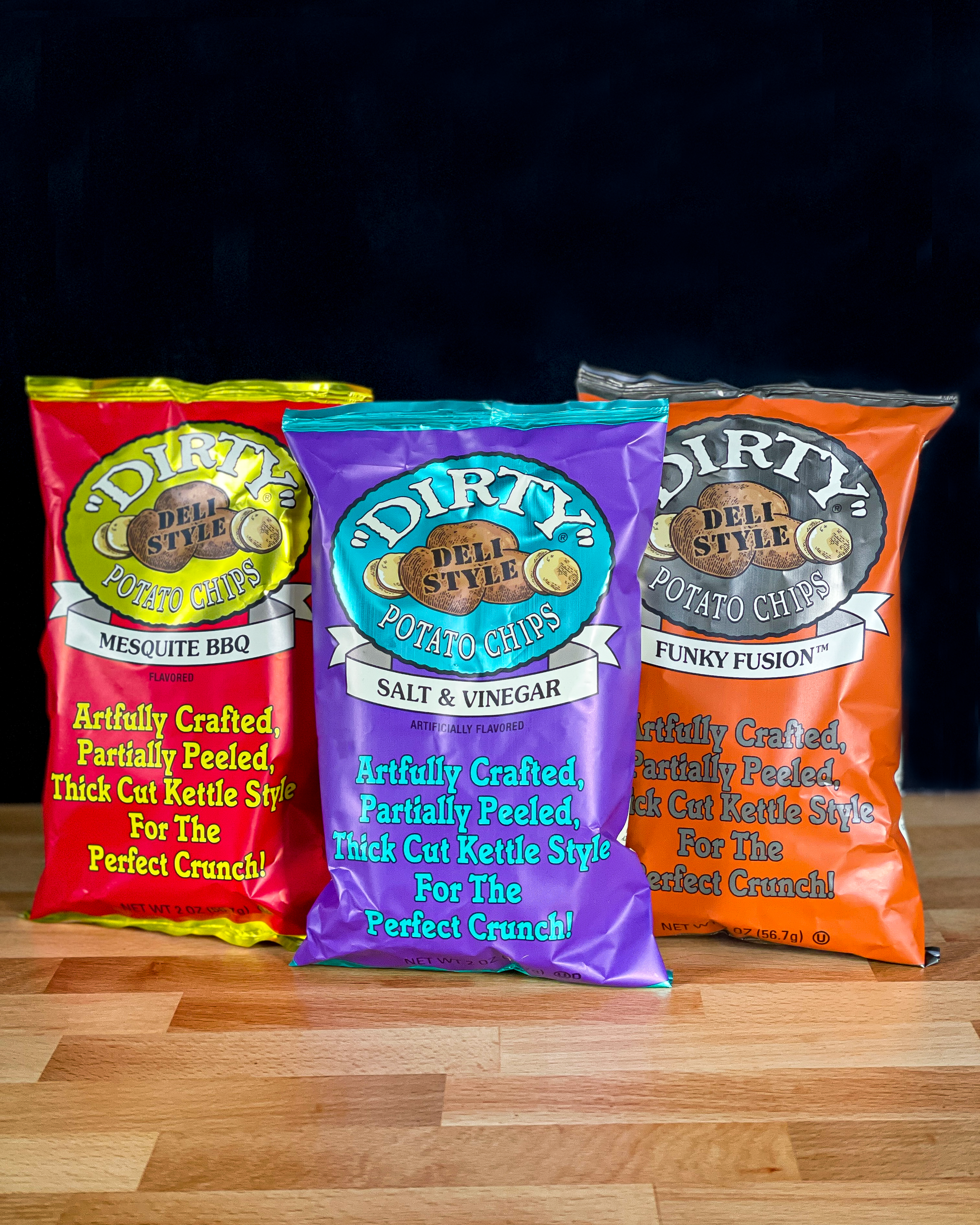 Three bags of Dirty Potato Chips in different flavors on a wooden surface with a black background.