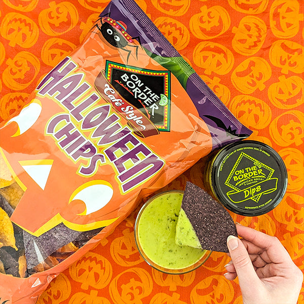 Halloween-themed tortilla chips packaging with a hand holding a chip and dip on an orange background with pumpkin designs.