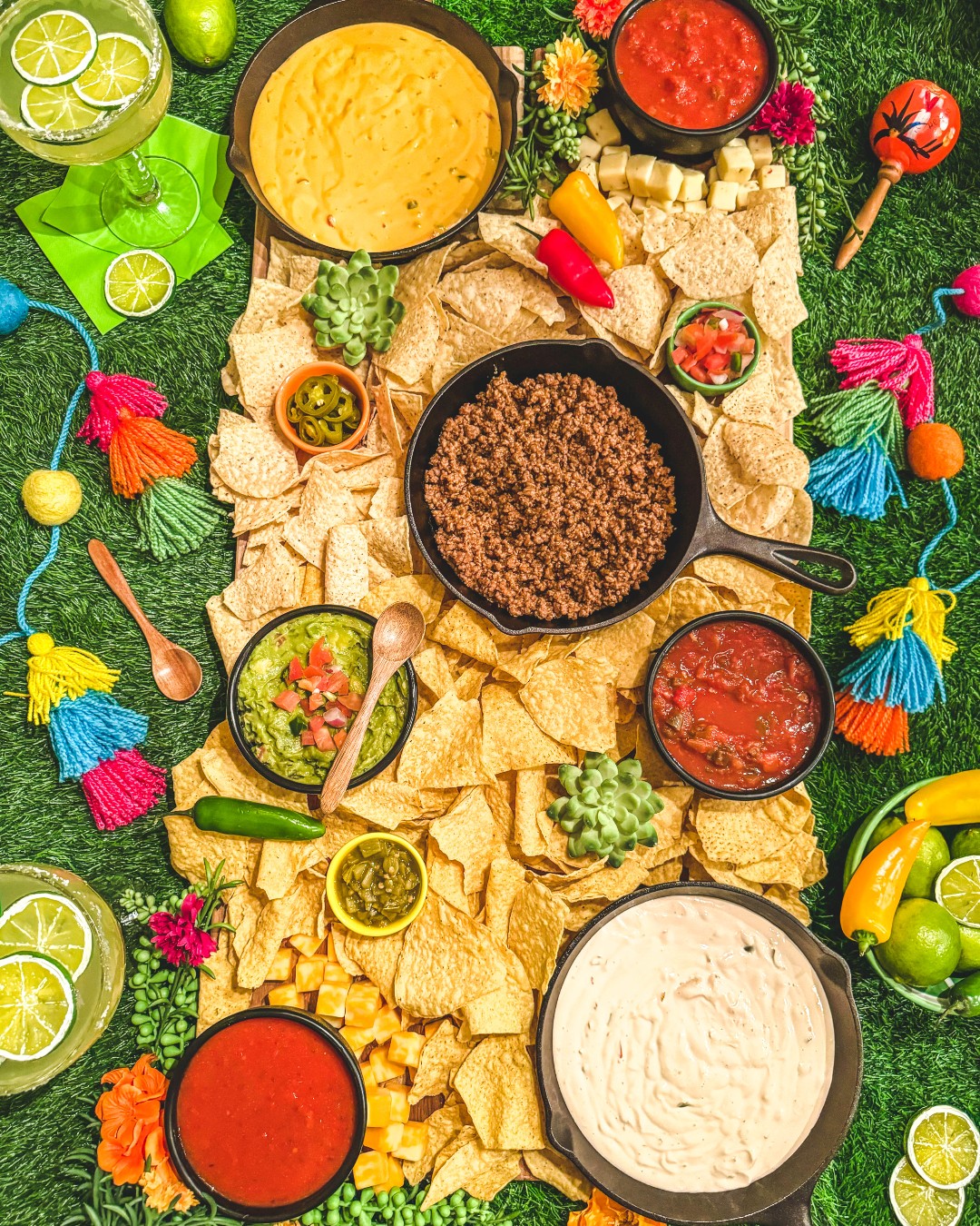 Assorted Mexican food including tortilla chips, ground beef, and various dips on a green surface with decorative items.