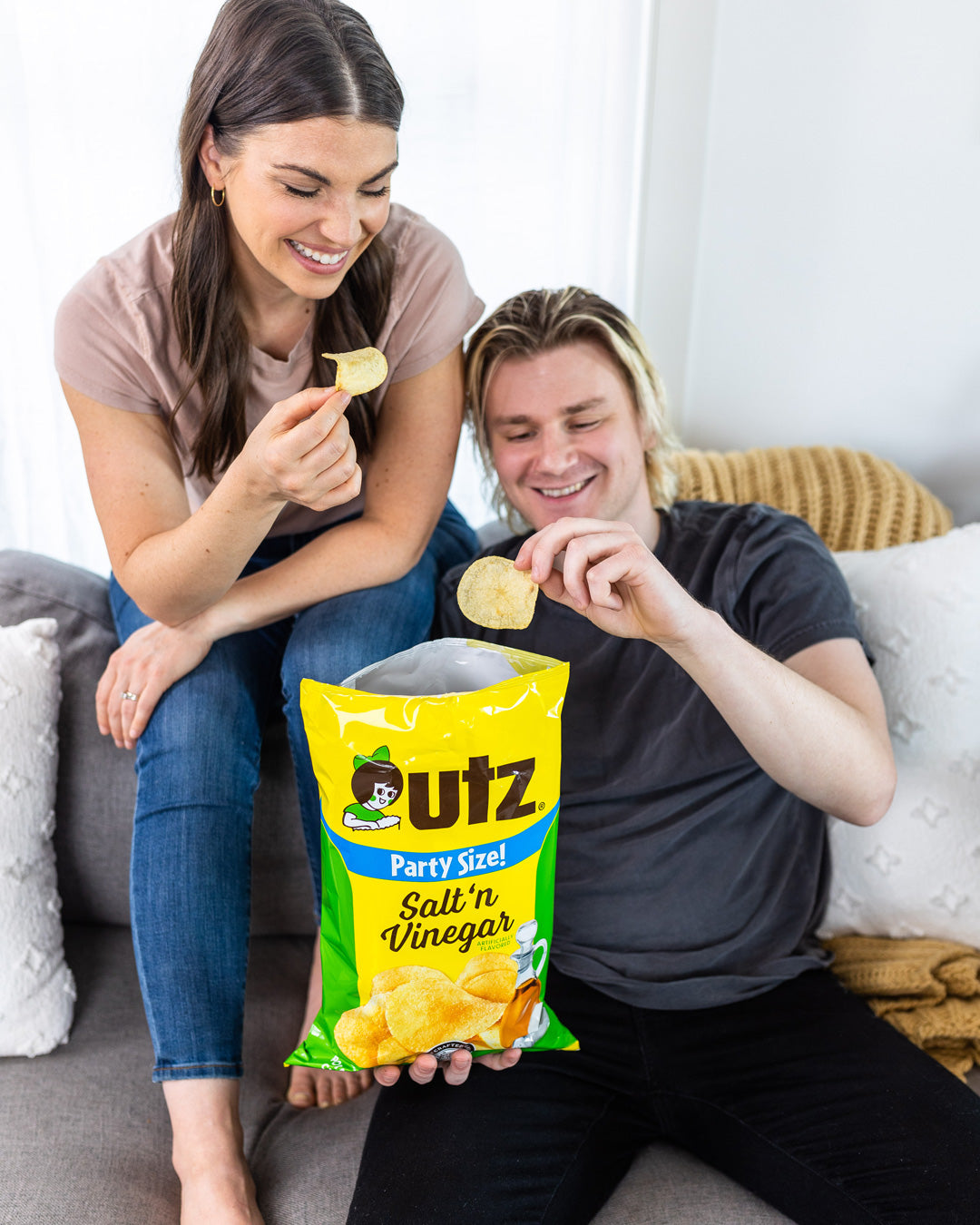 Two people sitting on a couch eating Utz salt 'n vinegar chips from a large bag.